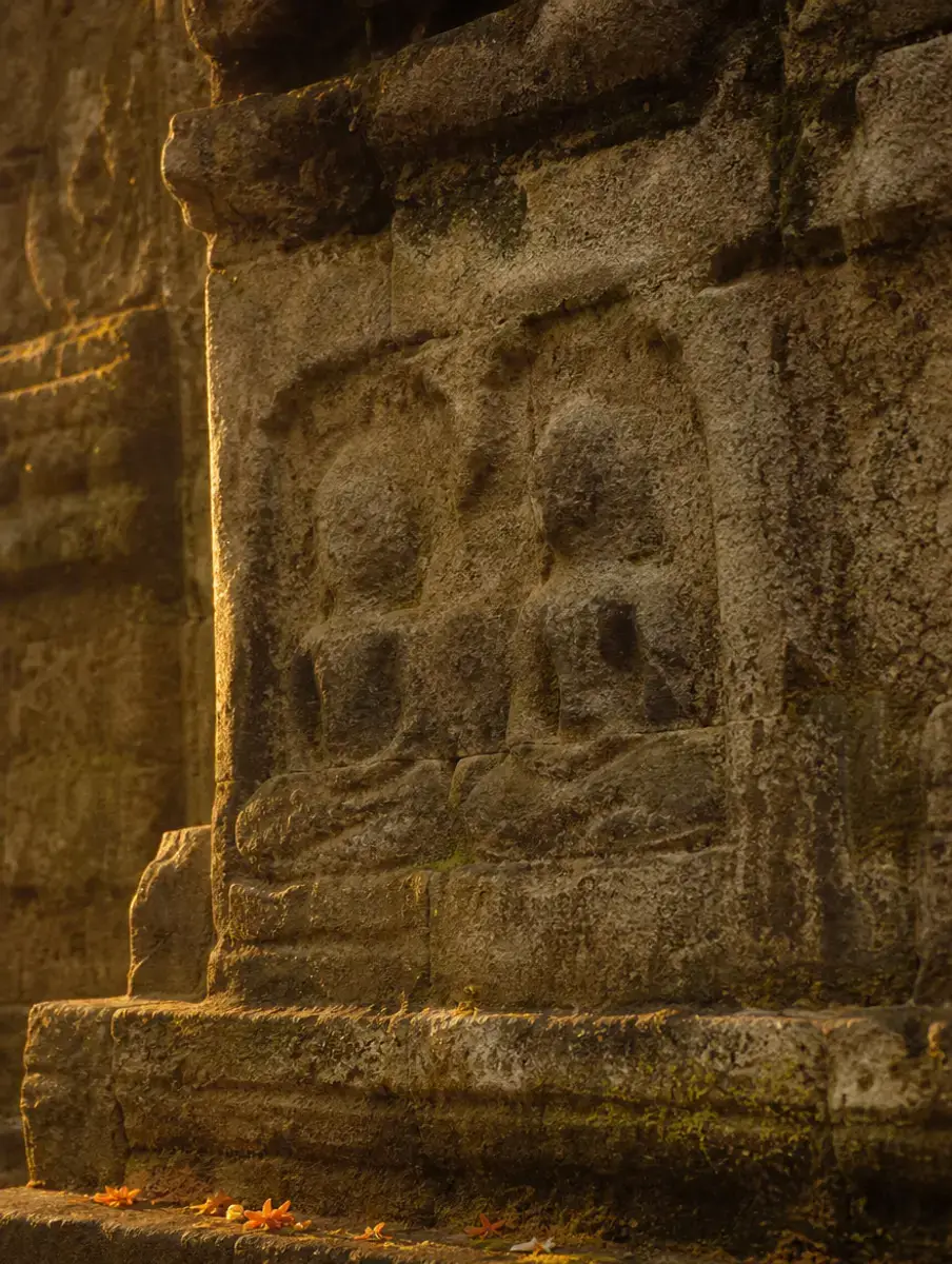A weathered sandstone temple wall with worn carvings of seated figures, lit by low golden light.