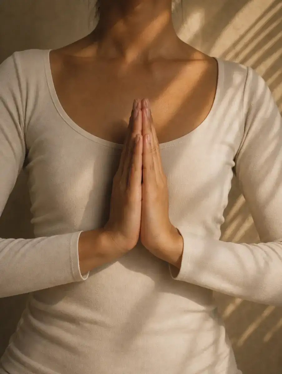 A faceless close crop of hands pressed together in anjali mudra at the centre of the chest, with palm-leaf shadows on a plaster wall.