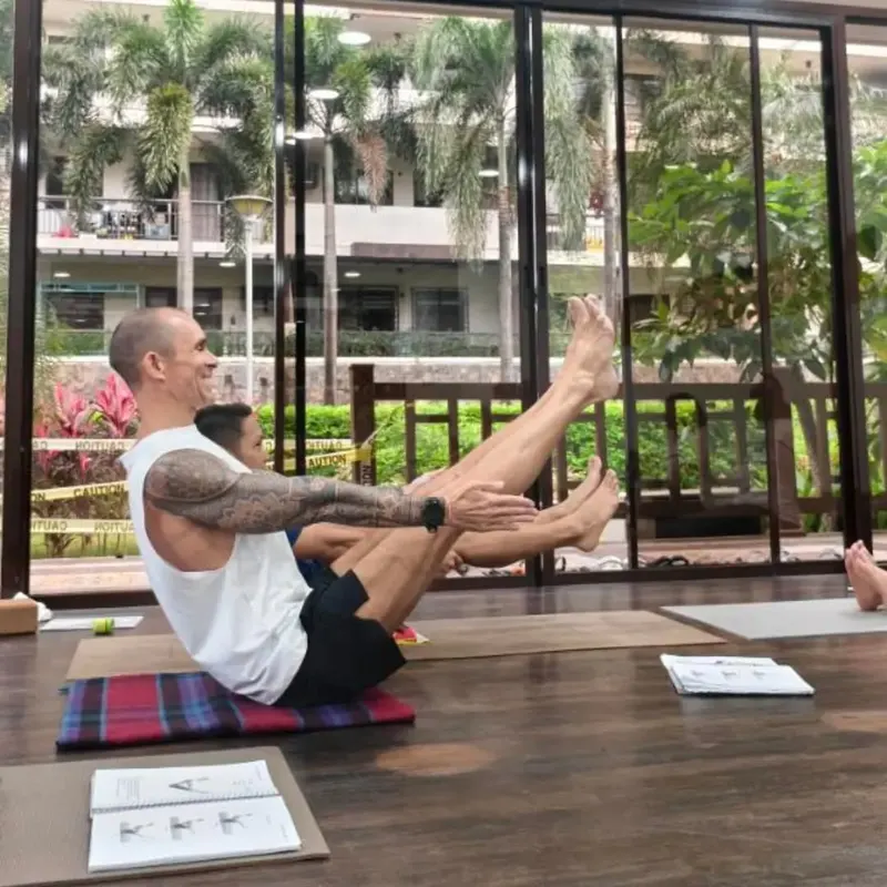 An AV108 teacher demonstrating boat pose in the window-lined practice room