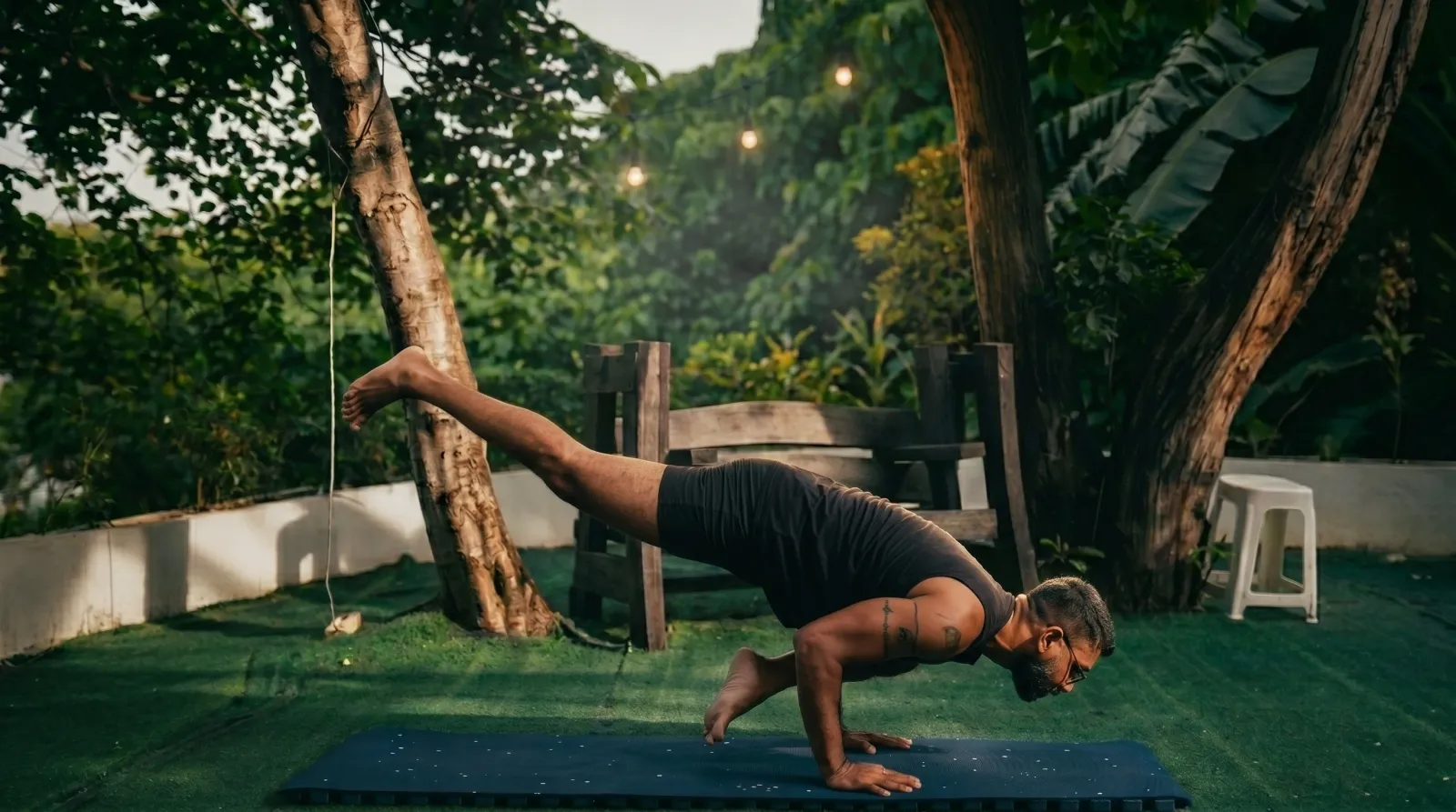 An Indian Yoga School PH teacher in a crow-pose variation under the garden trees