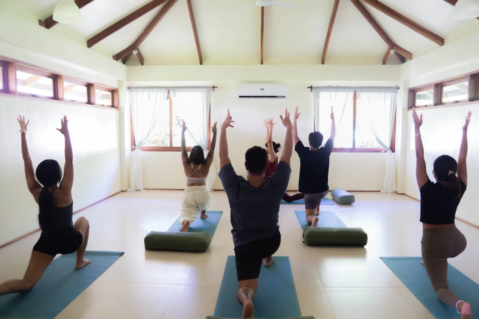 A class in low lunge with arms raised seen from behind at Little Shell Yoga Shala