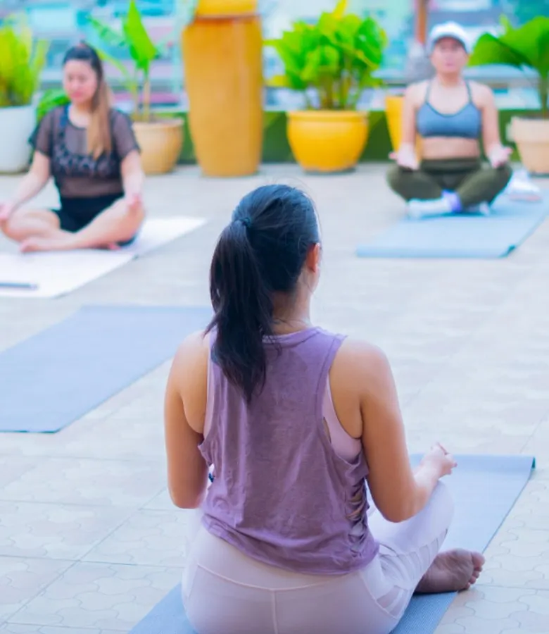 Seated meditation on mats on a rooftop deck with potted plants, seen from behind