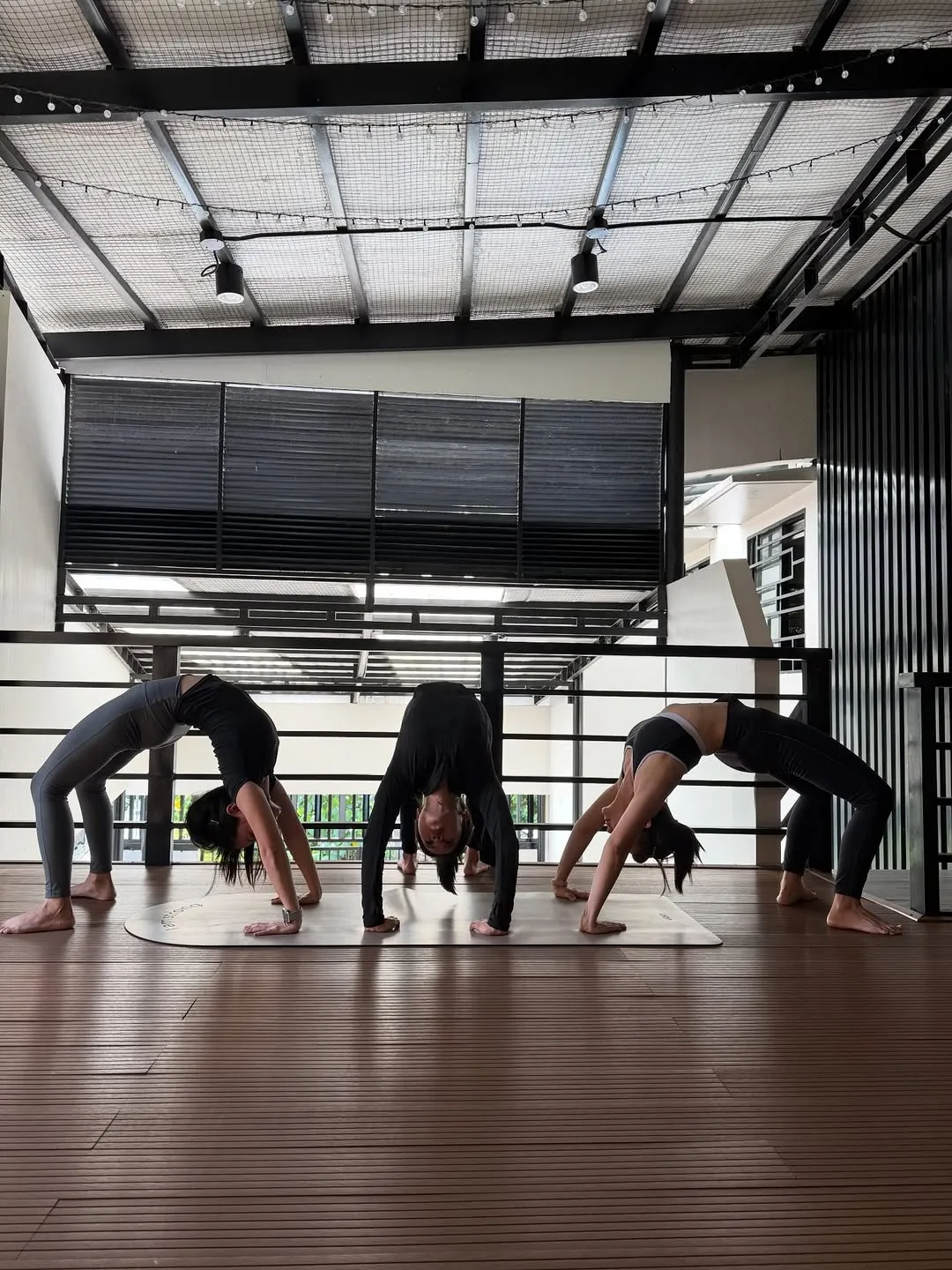 Students in wheel pose on a wooden floor under a high metal roof