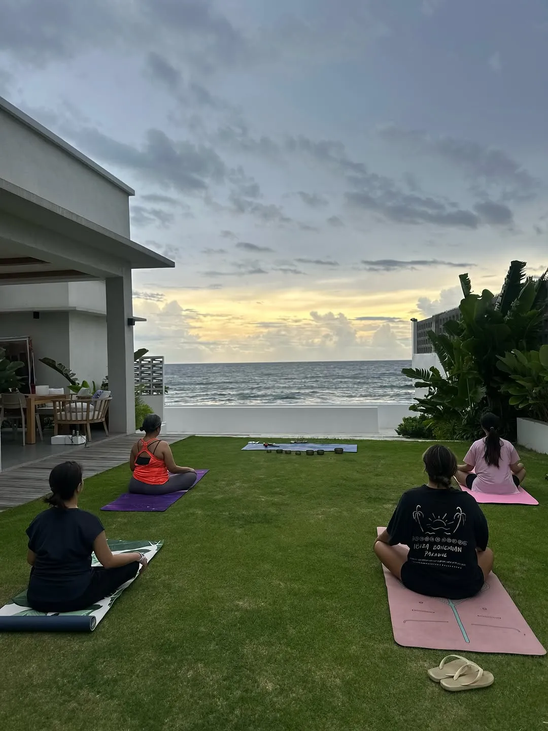 Seated students on mats on a lawn facing the sea at dusk