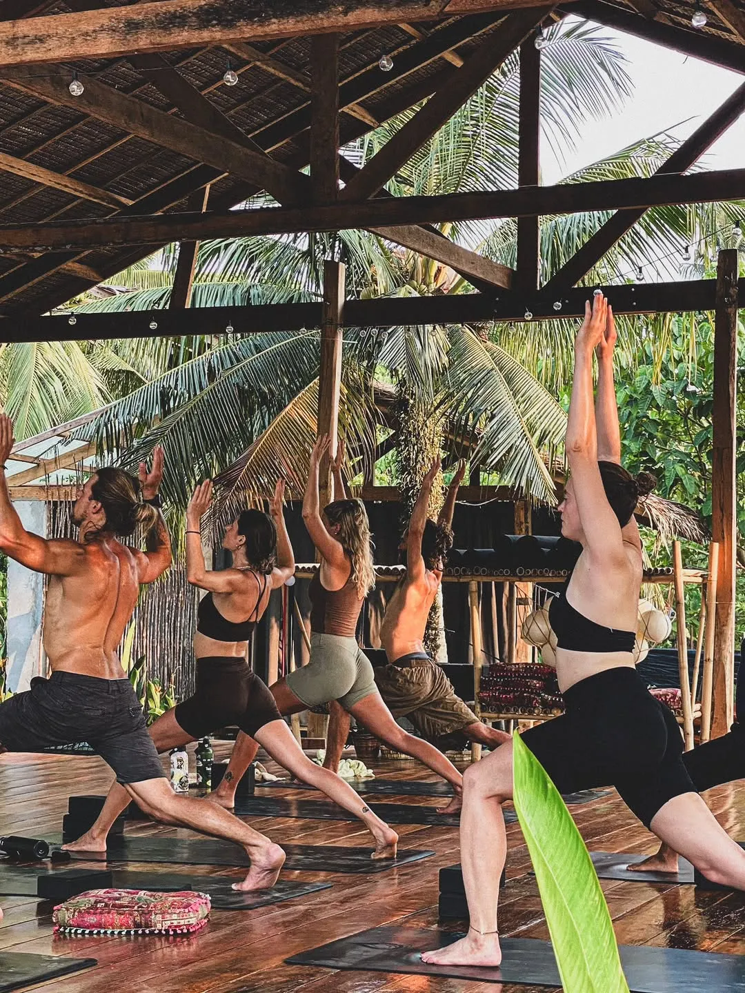 Class in a warrior pose in an open-air wooden shala surrounded by palms